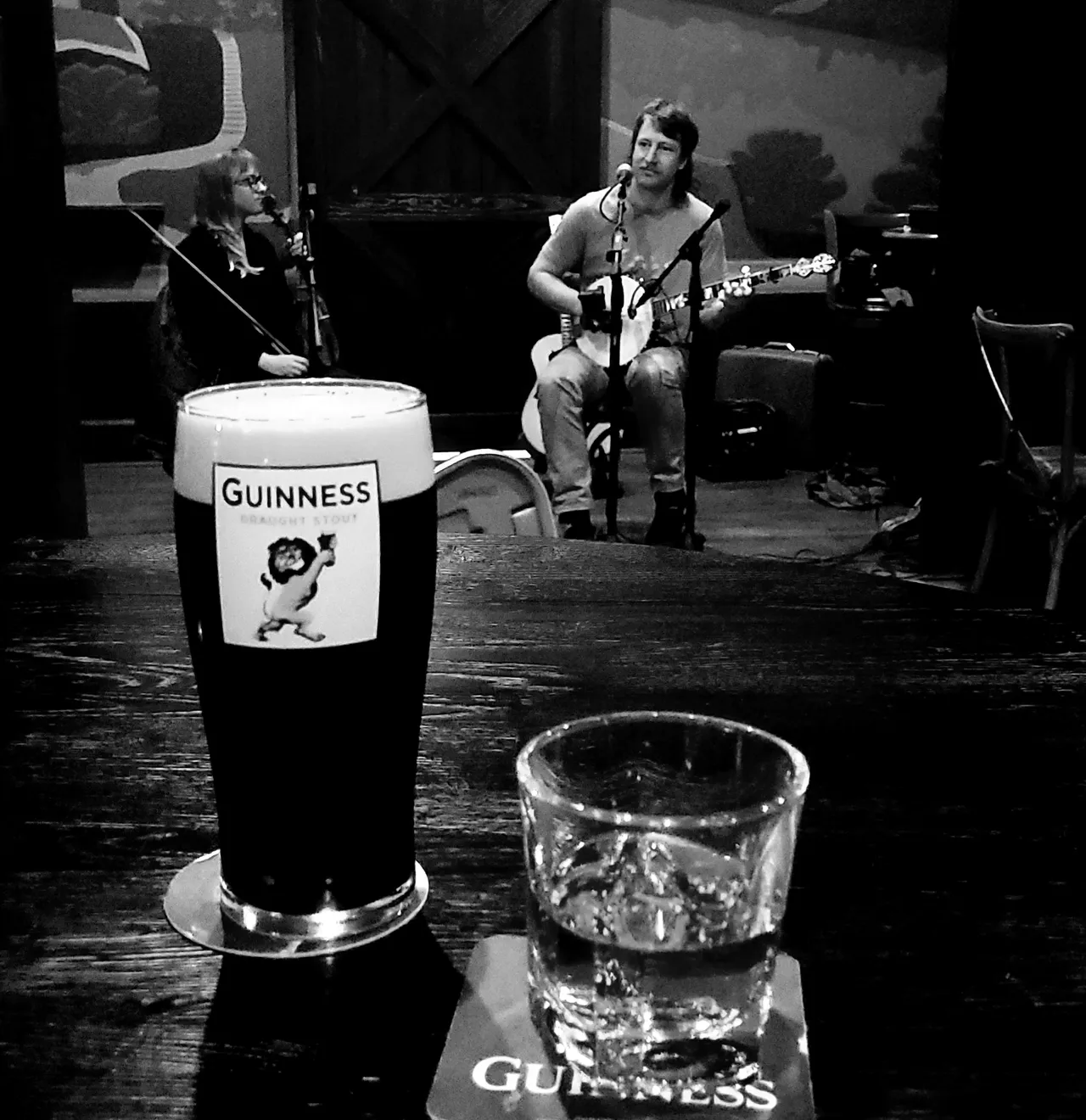 A Full glass of Guinness, a glass of clear liquor on a table in front of a banjo player and violinist.