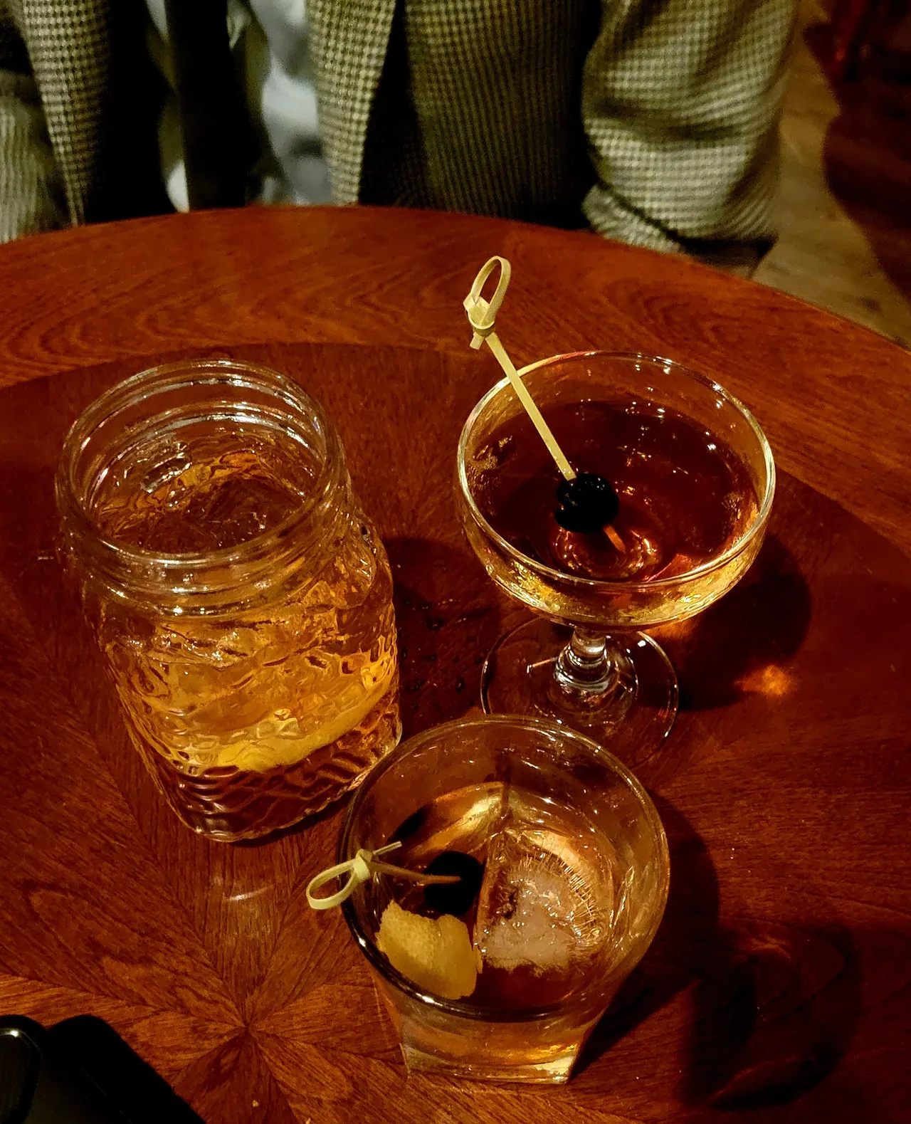 3 mixed drinks on a table. A mason Jar with a lemon in it, a stemmed glass with an olive on a stick, and a rocks glass with ice, olive and lemon.